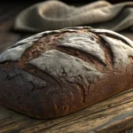 A close-up of a comically hard, misshapen loaf of homemade bread on a cutting board, next to a small...